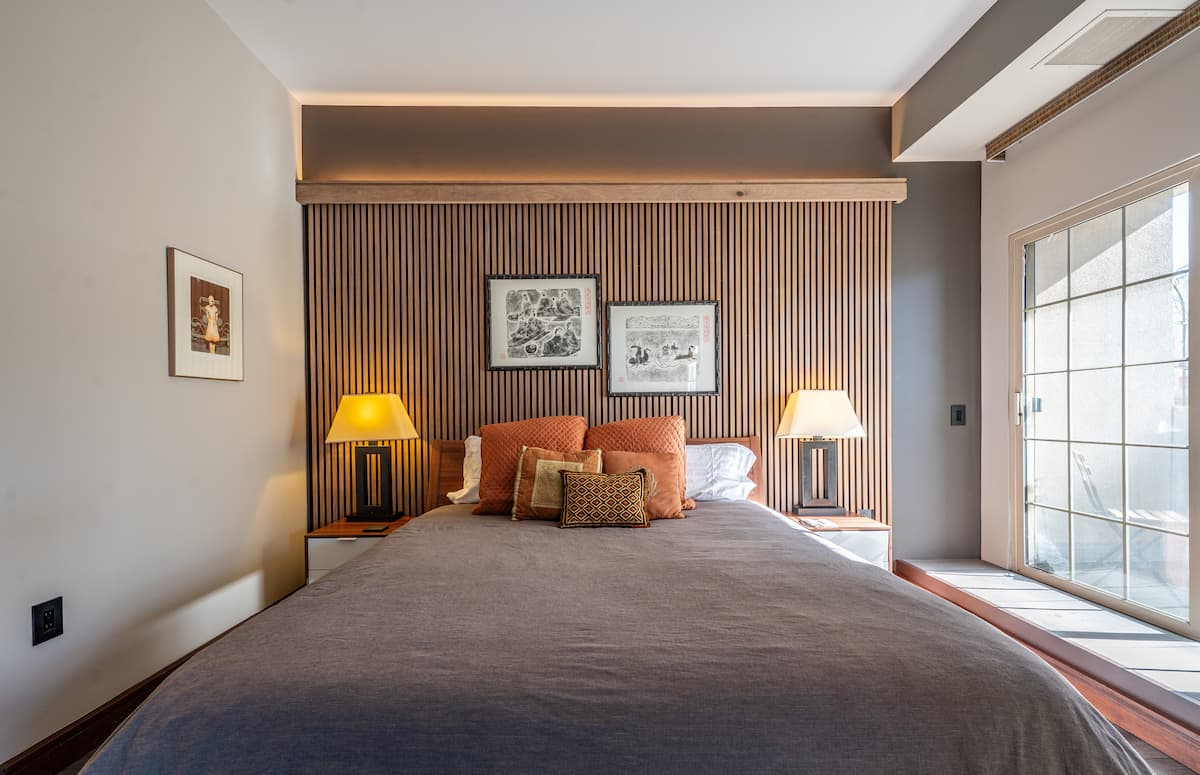 Bedroom with a king bed against a slatted-wood headboard wall, brass nightstand lamp, and a sliding glass door to the balcony.